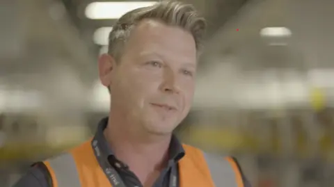 Amazon Wiktor Sierko has fair hair, with a quiff at the front. He has blue eyes and is wearing a grey shirt, lanyard and orange hi-vis vest.