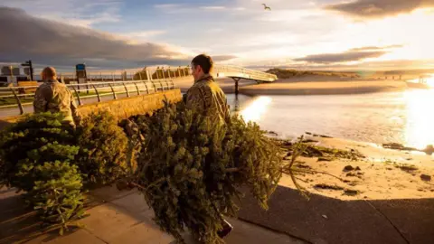 RAF Lossiemouth RAF personnel in camouflage carrying Christmas trees, with a bridge and water and sand dunes in the background, and a gull flying across a bright skyline.