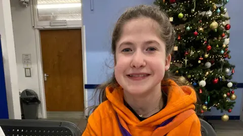 A girl with brown hair wearing an orange hoodie and neckerchief smiles at the camera. There is a Christmas tree behind her.