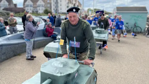 A man pushing a replica of a war tank on top of a shopping trolley. There is a group of people behind him doing the same; they are carrying flags, buckets and wearing t-shirts with the Motor Neurone Disease Association logo on it.