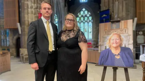 A smartly dressed man and woman stand together at the front of a church next to a picture on a stand of a blonde smiling woman dressed in blue