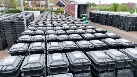Aerial view of thousands of black bins in a car park, with a lorry in the background and a forklift truck carrying bins around.