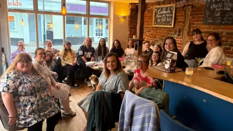 Lauren Emms A group of women sit within a pub in a circle. They are turned so they are looking at the camera and smiling. 