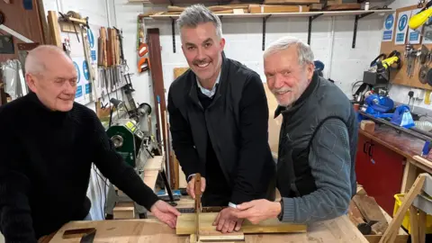 BBC/RICHARD EDWARDS This picture shows elected mayor David Skaith getting a woodwork lesson at York Mens Shed. Mr Skaith is sawing through a block of wood. Shed members Stuart Crossland and Peter Doherty are helping him.