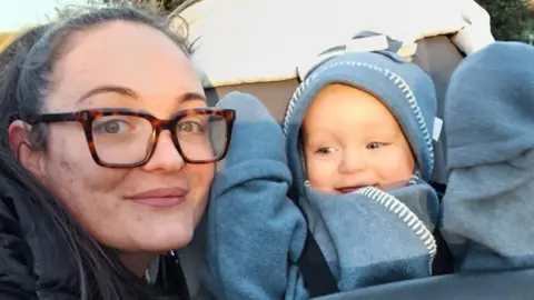Supplied A selfie- style image with Angela with her son Kai in a stroller. Angela hair brown hair held back in a ponytail and wears glasses. Kai is in a blue sleeper suit.