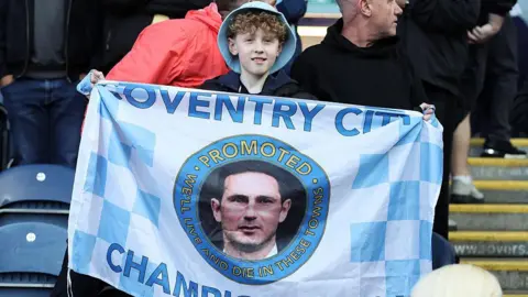 Getty Images A boy in a blue bucket hat holds up a blue and white Coventry City flag with the face of manager Frank Lampard in the middle