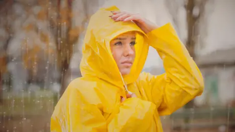 Getty A woman in a yellow raincoat looks worried as rain falls on her