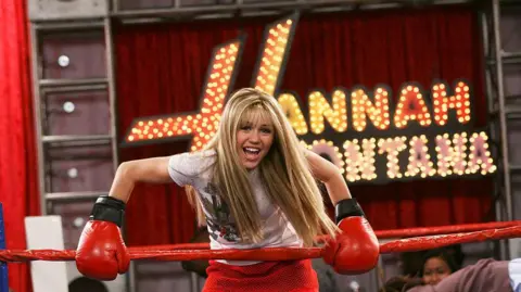 Getty Images Miley is wearing a long blond haired wig. She is in a red boxing ring holding the edge clad in red boxing gloves. Big letters with lights behind her read Hannah Montana. 