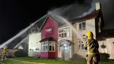 Two firefighters spraying water canons over the red and white Toby Carvery building. It is still dark