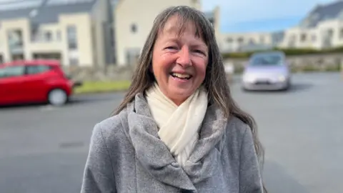 A women with long greyish hair smiling at the camera. She has a white scarf and a grey coat on