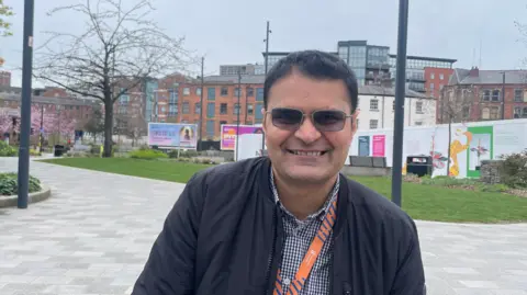 Sofía Luis-Hobbs/BBC A man with tinted sunglasses and brunette hair smiles at the camera. He wears a blue and white chequered shirt beneath a black zip-up bomber jacket. Behind him there are buildings in the background as well as some greenery in front of a boarded off building area.