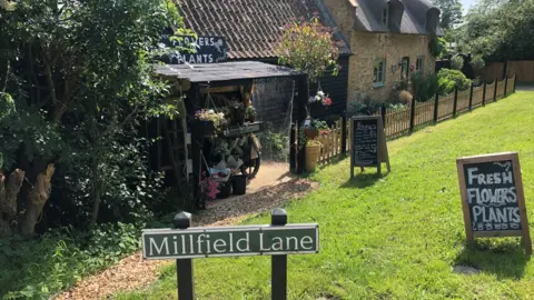 BBC/John Devine A flower stall outside a cottage behind a sign reading Millfield Lane