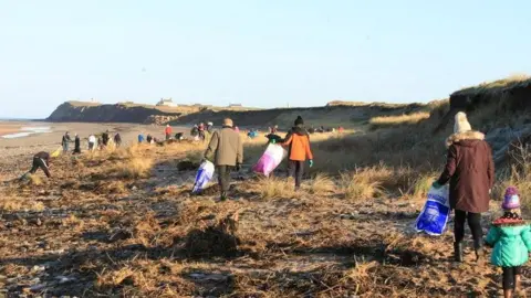 BEACH BUDDIES Wide shot of Ballaugh Beach - you can see the headland in the background. There are about 100 people scattered across the beach, picking up rubbish.