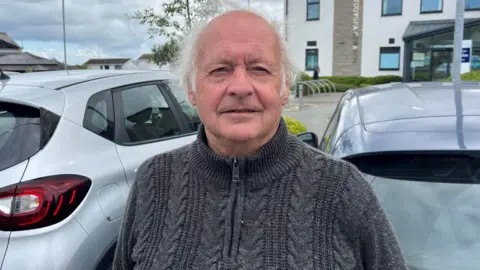 A man with grey hair, stood outside in a car park. He is looking directly at the camera. He is wearing a dark grey knitted quarter zip jumper, with a high neck.