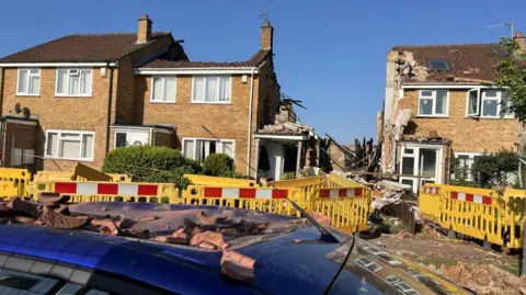 A row of yellow brick houses with a gap in the middle filled with debris. The roof is missing from one of the houses and another is badly damaged. Yellow safety barriers surround the site.