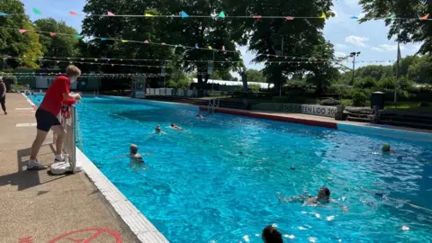 Tom Jackson/BBC People swimming in an outdoor pool. There is a lifeguard in a red T-shirt and black shorts on duty. Overhead multi-coloured bunting has been strung across the pool.