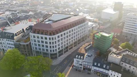 BBC Jersey's government offices seen from the air. The mainly white building is next to a road and surrounded by other buildings. The tops of two trees can be seen in the lower left hand corner.