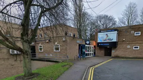 Chester-le-Street Leisure Centre: a two-storey brick building with a glass door entrance at its centre with, at a right angle, another part of the building featuring an overhanging window filled by an image of a woman doing yoga