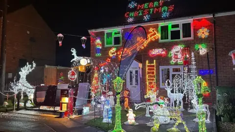 The house is covered with numerous lights and decorations with others in the front of the garden. The decorations include reindeers covered in lights, Santa's sleigh and snowmen.