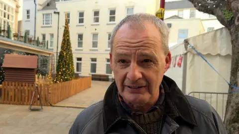 Ian Barette looking at the camera, wearing a wax jacket and knit jumper. He has short grey hair. He is standing in Jersey's Royal Square with trees and paving visible behind him. 