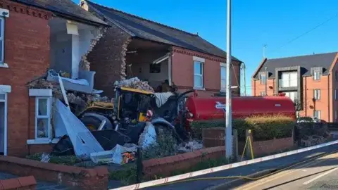 A tractor pulling a red tanker crashed into two terraced houses. The front of the houses is totally destroyed.