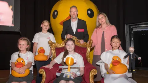 Four girls are sitting on chairs, wearing white t-shirts and holding yellow rubber ducks. They are smiling next to Alex Horne who is wearing a white shirt and a black blazer. He is standing in front of a giant yellow rubber duck. To the right of the girls is a woman with long blonde hair wearing a pink jacket and a dress.