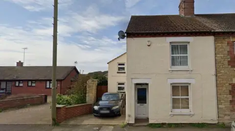 Google Small two-storey cream-rendered end-of-terrace house with one window on each floor and a white-coloured door set back from the front facade. A car is parked in the drive to the left and there is a two-storey extension visible behind the car. A brick-built bungalow with a gravel drive is visible to the left.