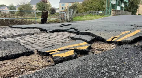 Harriet Heywood/BBC Images shows Mill Lane road and chunks of tarmac with yellow lines on them broken up with the cobbles underneath exposed.
