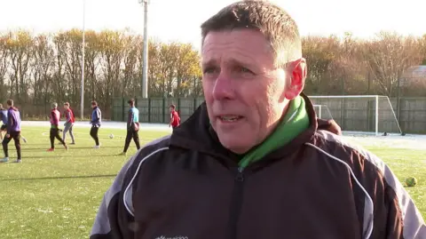 David Dodds is standing on the pitch with players in action behind him. He is a middle-aged man wearing a grey sports jacket with a green scarf. He looks cold. 