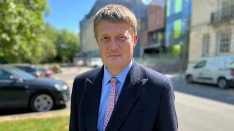 Richard Clewer looks into the camera. He wears a suit with shirt and tie - in the background, the blurred Wiltshire council HQ on a sunny day