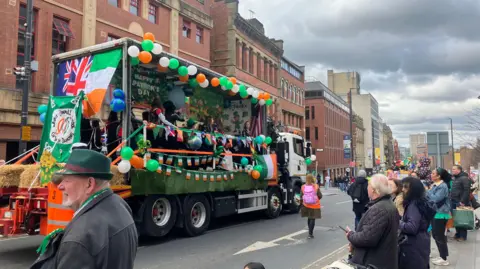 An open-sided truck decorated with flags and balloons drives down The Headrow in Leeds. On the truck are dancers in green, orange and white.