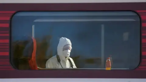 Joe Giddens/PA The head and shoulder of a police forensics officer is visible through a train carriage window. Their white hood is over their head, and they have a white mask over their nose and mouth.