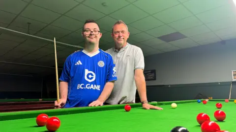 Dylan and his dad stand together at a green snooker table in a club. Dylan is wearing a blue sports jersey with the words: “BC.GAME”. He is holding a cue stick upright beside the table edge. His dad stands slightly behind with one hand resting on the table. Several red balls are scattered across the felt in the foreground, with a black ball near the bottom edge and a white cue ball and coloured balls visible further back.