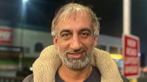 Elizabeth Baines / BBC A man with grey hair wears a fleece lined jacket. Behind him, road signs are blurred.
