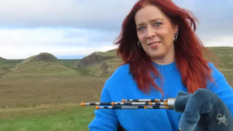 Highlights PR Head and shoulder shot of a smiling woman with red hair, wearing a blue jumper and holding a set of Northumbrian pipes. Behind her can be viewed moorland and rolling hills