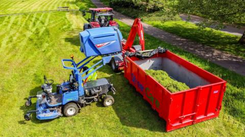 A green field upon which is a red tractor and skip. There is also a blue vehicle that can help deposit material into the skip alongside it.