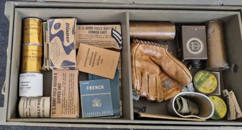 Cathy Roberts A box of US Army rations and other equipment, including canned food, a French dictionary, and a baseball catcher's mitt.