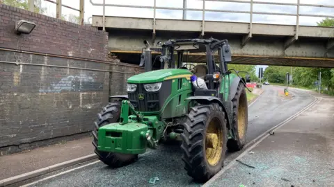 A green John Deere tractor has a badly damaged roof and wing mirrors. It is on a road under a bridge and there is debris and shattered glass on the road and pavement.