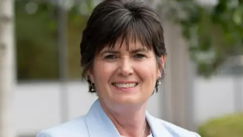 A photo of a woman with short dark brown hair smiling into the camera. She has got a blue blazer on and earrings. 