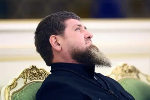 Reuters Ramzan Kadyrov em uma cadeira ornamentada com uma barba longa e cheia olhando ligeiramente para cima com uma expressão contemplativa. Ele está vestindo uma camisa escura com gola alta. Em volta do pescoço há um colar de contas pretas ou um colar de contas.