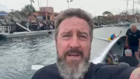 Andy Merry taking a selfie off the side of a rowing boat with other rowers behind him in a harbour.  He has a thick greying beard.