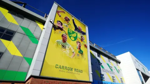 The outside of Carrow Road football stadium in Norwich. There is yellow and green branding across the building.