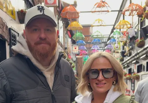 A man and woman are looking into the camera. the man on the left is wearing a white baseball cap, a white hoody and a grey zip up jacket. He has a ginger beard. The woman has shoulder-length blond hair, a grey hoody, green coat and and tortoiseshell glasses. Behind them is a number of colours umbrellas hanging from a metal grid. 
