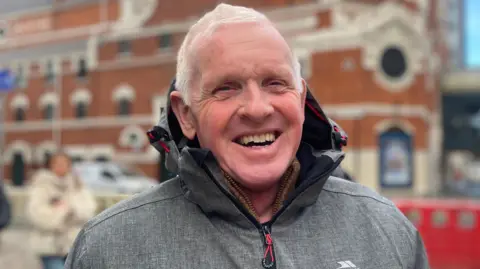 Gregory Gault wears a brown jacket. He has grey hair and is smiling. Behind him is the Grand Opera House, Belfast.