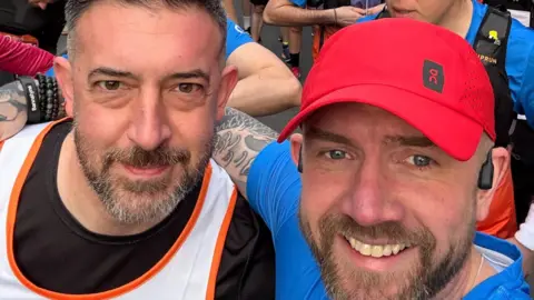 Two smiling men with beards in a crowd of other people, with the person on the right wearing a red cap and blue top and the man on the left a black top and white vest
