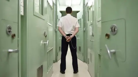 A male prison officer standing in a prison, in uniform, with his hands behind his back and his back to the camera. He's standing in a narrow corridor between two rows of cells with light green doors.