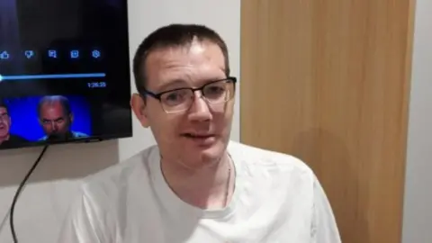 Brainkind Neurological Centre Karl is wearing a white top and has dark hair and glasses and is stood in front of a wooden door and television