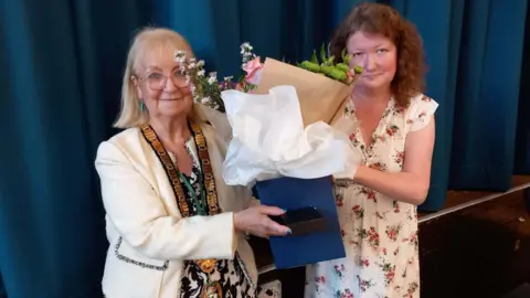 BBC Councillor Sue Eacock, left, pictured with Councillor Joanna Kane when she became the new Mayor of Redditch - Councillor Eacock is wearing a white jacket with a large gold chain around her neck. She is being handed flowers to mark the occasion.