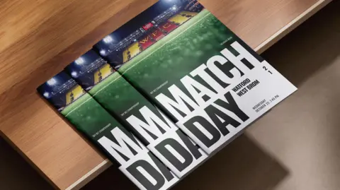 Watford FC A stack of football programmes rests on a wooden surface. The covers feature a football stadium at the top with bright floodlights and yellow and red seating. Below the stadium photo, large bold text reads “MATCH DAY".