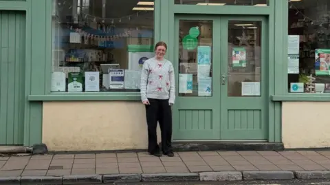 A wide shot of Madison Oman standing outside her workplace. She has tied back brown hair and is wearing a grey Christmas jumper and dark trousers.